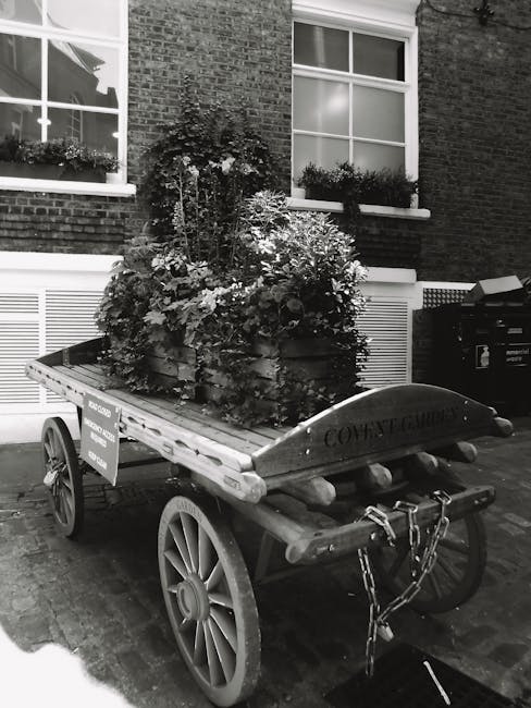 A rustic wooden cart with large spoked wheels is parked on a brick-paved street in front of a brick building with large, double-hung sash windows that have ornate white frames. The cart is loaded with